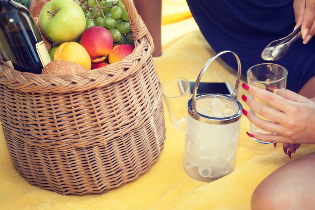 Close-up of a summer birthday picnic scene with a woven basket filled with wine, burgers, apples, peaches, and grapes placed on a yellow picnic blanket. A guest uses tongs to add ice from a clear bucket into a glass, with another empty glass and a smartphone nearby. The setup highlights a relaxed and refreshing picnic atmosphere ideal for warm-weather celebrations.