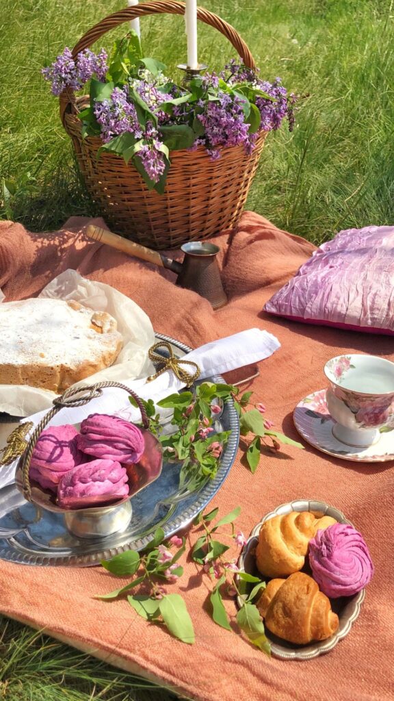 Romantic spring birthday picnic setup on a peach blanket in the grass, featuring a wicker basket filled with fresh lilac flowers and white candles. The scene includes a silver tray with purple meringue pastries, croissants, a powdered cake, a vintage teacup, and folded napkins, surrounded by blooming branches and soft sunlight—perfect for a charming outdoor tea picnic celebration.