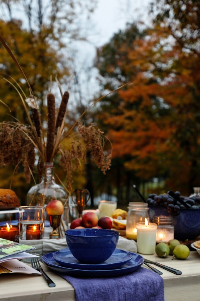 Elegant fall-themed birthday picnic table set outdoors, featuring deep blue ceramic dishes on a purple linen napkin, surrounded by candles, apples, grapes, figs, and rustic glass jars. A centerpiece of dried wildflowers in a clear vase enhances the autumn atmosphere, with golden leaves and trees in the background creating a warm and cozy seasonal setting perfect for an intimate outdoor celebration.