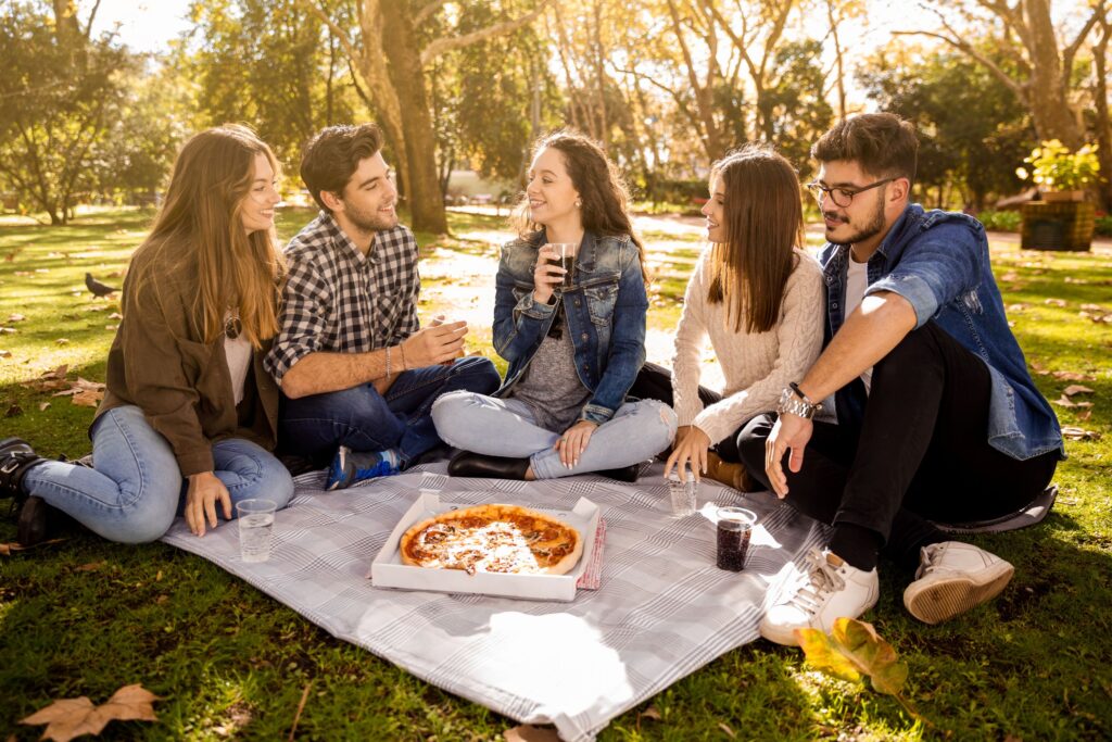 Group of five friends enjoying a casual birthday picnic in a sunny park, sitting on a gray checkered blanket with a box of pizza and plastic cups filled with soda. Surrounded by green grass and autumn leaves, the group chats and laughs under the shade of tall trees, capturing a relaxed and youthful outdoor celebration vibe.