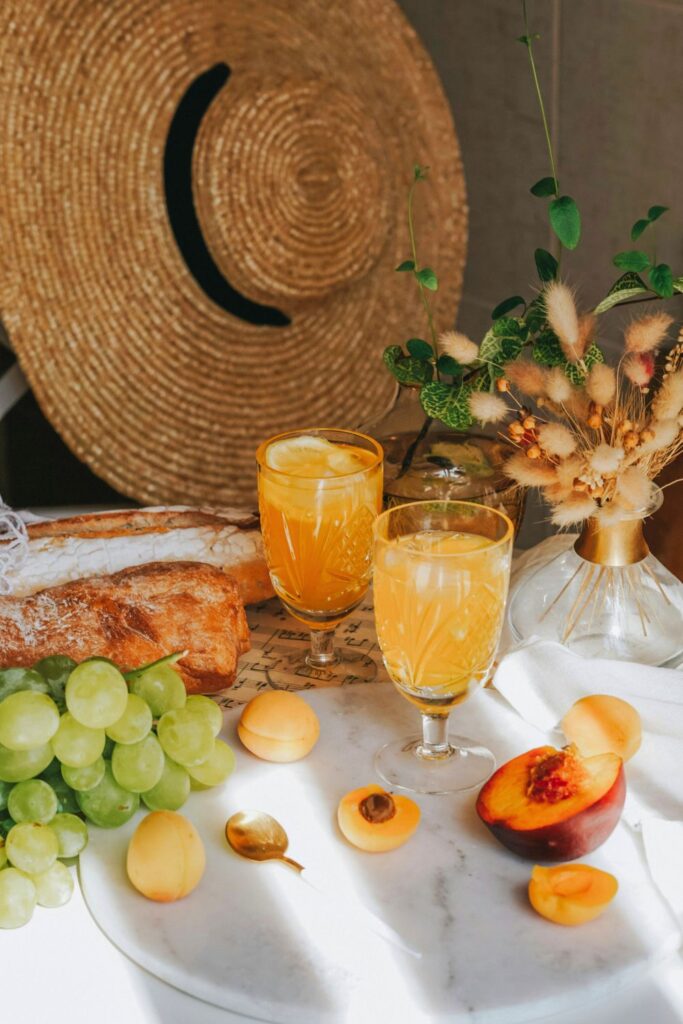 Fresh summer picnic scene with golden-hued iced drinks in vintage glasses, surrounded by sliced peach, apricots, green grapes, crusty bread, and dried floral decor, creating a warm, rustic, and inviting tabletop setting.