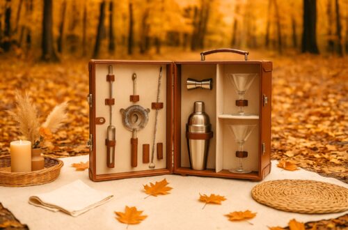 Luxury cocktail picnic set displayed in an autumn forest surrounded by golden and orange leaves. The tan leather case is open on a cream blanket, showing neatly organized stainless steel bar tools including a shaker, jigger, strainer, muddler, and spoon, along with two martini glasses secured inside. Boho picnic accents such as a wicker tray with a candle, dried pampas grass in a vase, folded napkins, and a woven placemat complete the cozy outdoor setup. Warm golden sunlight filters through the forest, highlighting the seasonal fall atmosphere.
