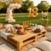 A beautifully arranged outdoor 18th birthday picnic setup in a green park during golden hour. The scene features a low wooden table covered with plates of sandwiches, fruit platters, and cupcakes, surrounded by beige cushions and soft blankets. A wicker basket and coolers sit nearby, while a white balloon arch and large gold “18” balloons serve as the decorative focal point. In the background, string lights and floral arrangements add a warm, elegant touch under the evening sunlight, creating a cozy, natural atmosphere perfect for a milestone celebration.