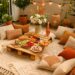photo of a boho-style balcony picnic setup in a small urban apartment. The balcony floor is covered with layered woven picnic blankets and colorful patterned rugs, surrounded by numerous decorative cushions in warm shades of terracotta, cream, mustard, and sage. A low wooden table holds an assortment of picnic food, including fresh fruit, pastries, and glasses of juice. The scene is styled with potted plants, hanging flowers, and rattan baskets, while lanterns and woven trays add cozy texture. Soft daylight highlights the mix of natural materials, creating a relaxed, vibrant, and inviting boho balcony decor perfect for an outdoor meal.