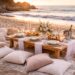 Luxury beach picnic setup at sunset with elegant wooden table, cushions, candles, floral centerpiece, and ocean view, showing a romantic outdoor picnic arrangement on a Malibu-style sandy beach.